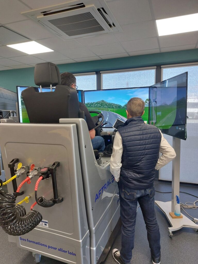 L'image montre deux hommes à l'intérieur d'une salle de formation, l'un conduisant un simulateur de véhicule avec plusieurs écrans affichant un paysage routier.