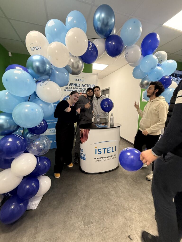 Un stand festif décoré de ballons bleu et blanc avec plusieurs personnes souriantes posant devant un panneau indiquant ISTELI, une école de gestion du transport et de la logistique.