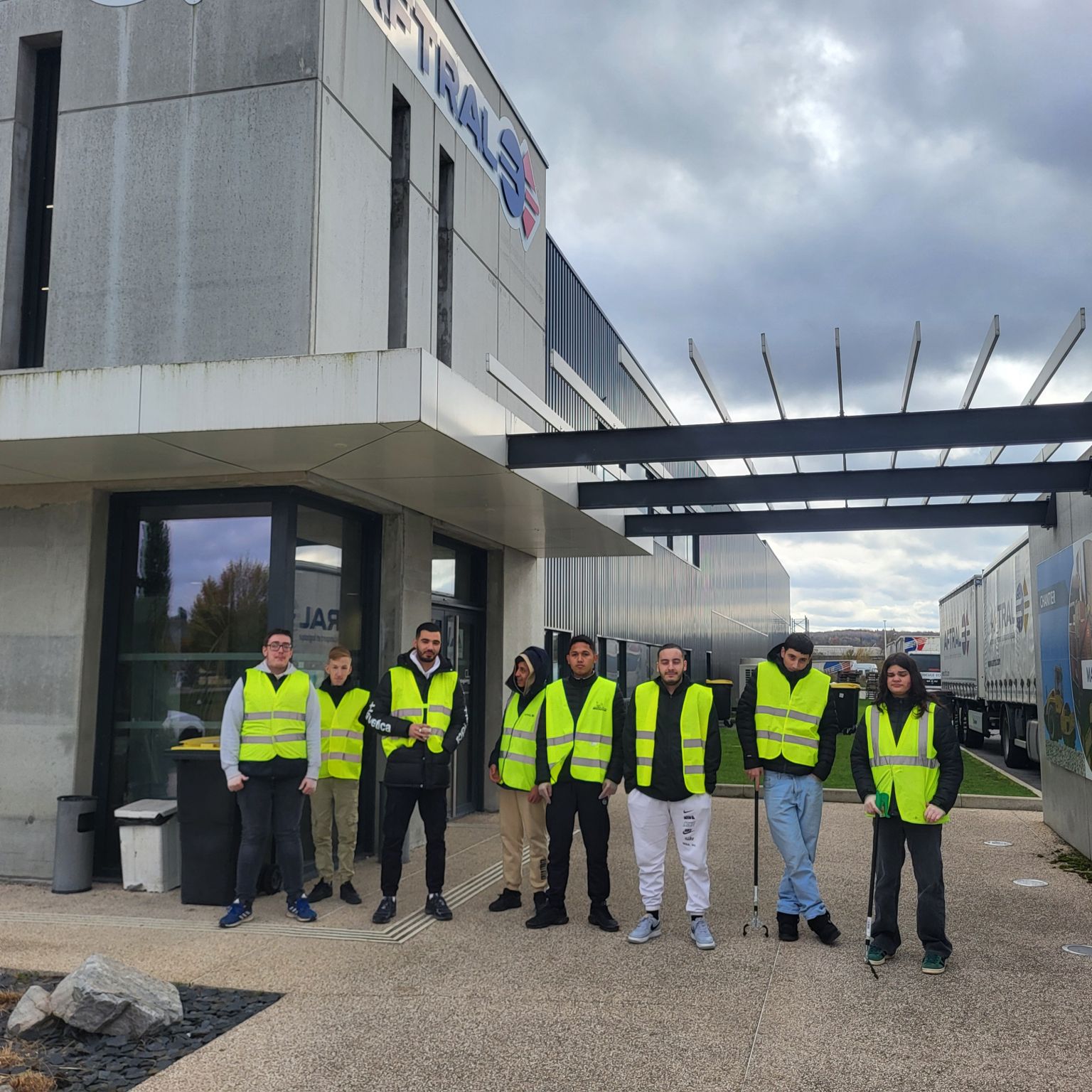 Un groupe de personnes portant des gilets de sécurité jaunes se tient devant un bâtiment moderne avec des camions stationnés à l'arrière-plan.