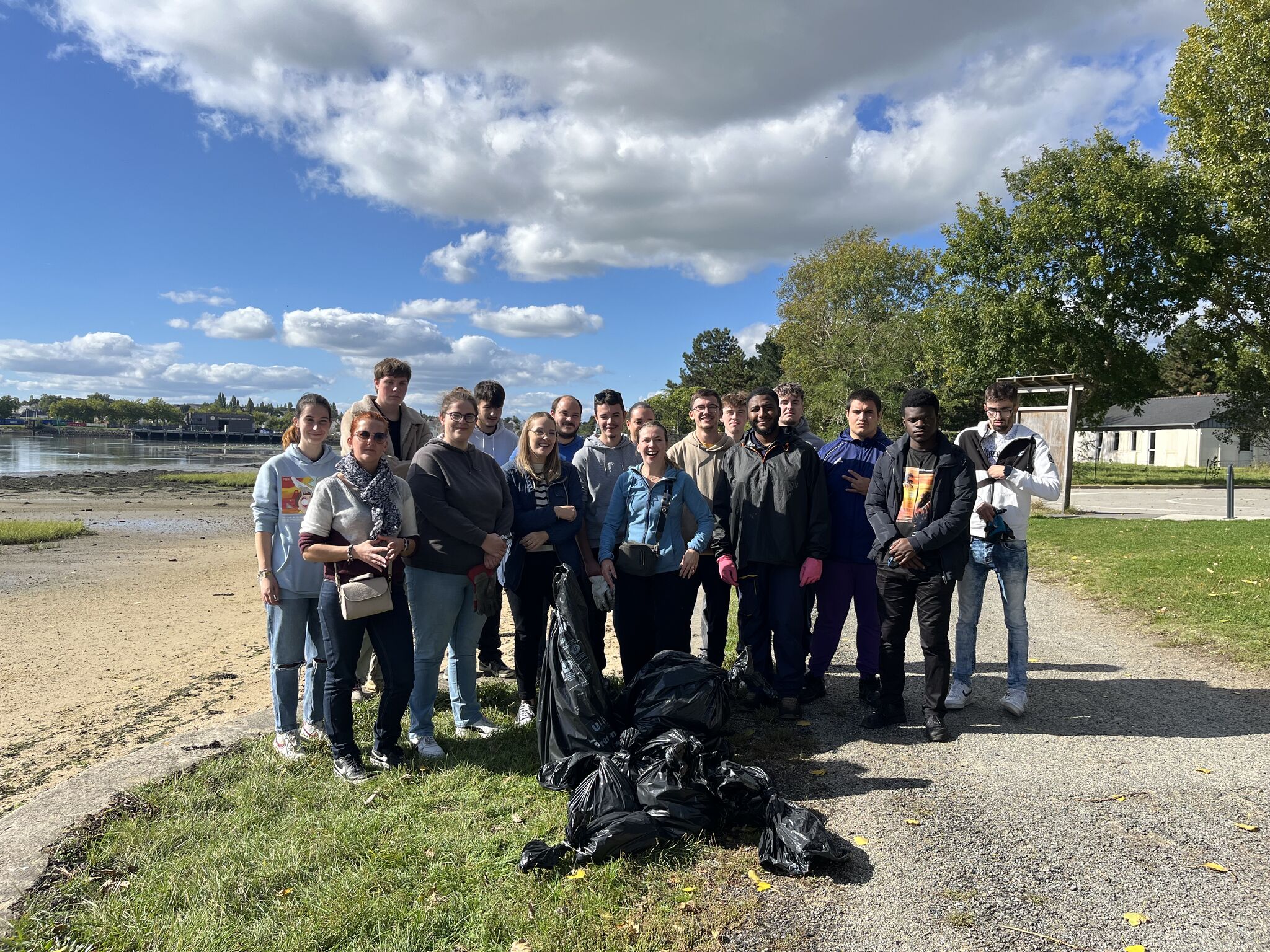 Un groupe de personnes se tient ensemble à l'extérieur, souriant, avec des sacs poubelles noirs au sol et un paysage de parc en arrière-plan.