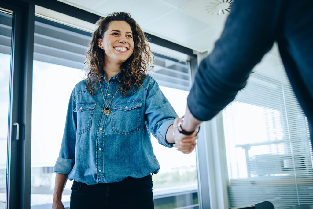 Une femme souriante, vêtue d'une chemise en denim, serre la main d'une autre personne dans un bureau moderne.