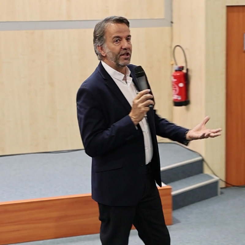 Un homme en costume foncé parle avec un microphone sur une scène devant un public dans une salle de conférence.