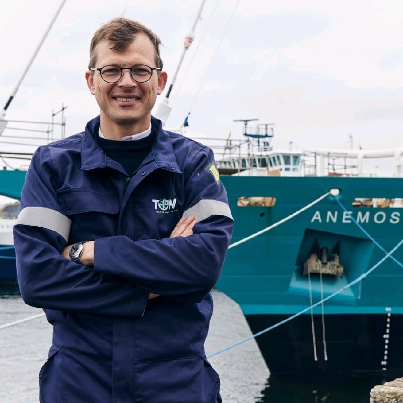 Un homme souriant en tenue de travail se tient devant un bateau de pêche bleu portant le nom "ANEMOS".