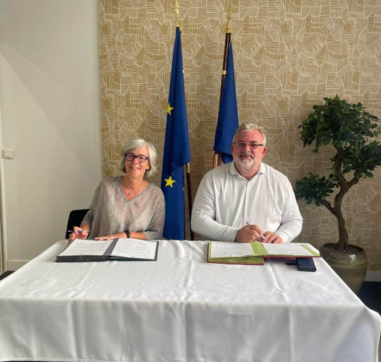 Deux personnes assises à une table signent des documents, avec des drapeaux et une plante verte en arrière-plan.