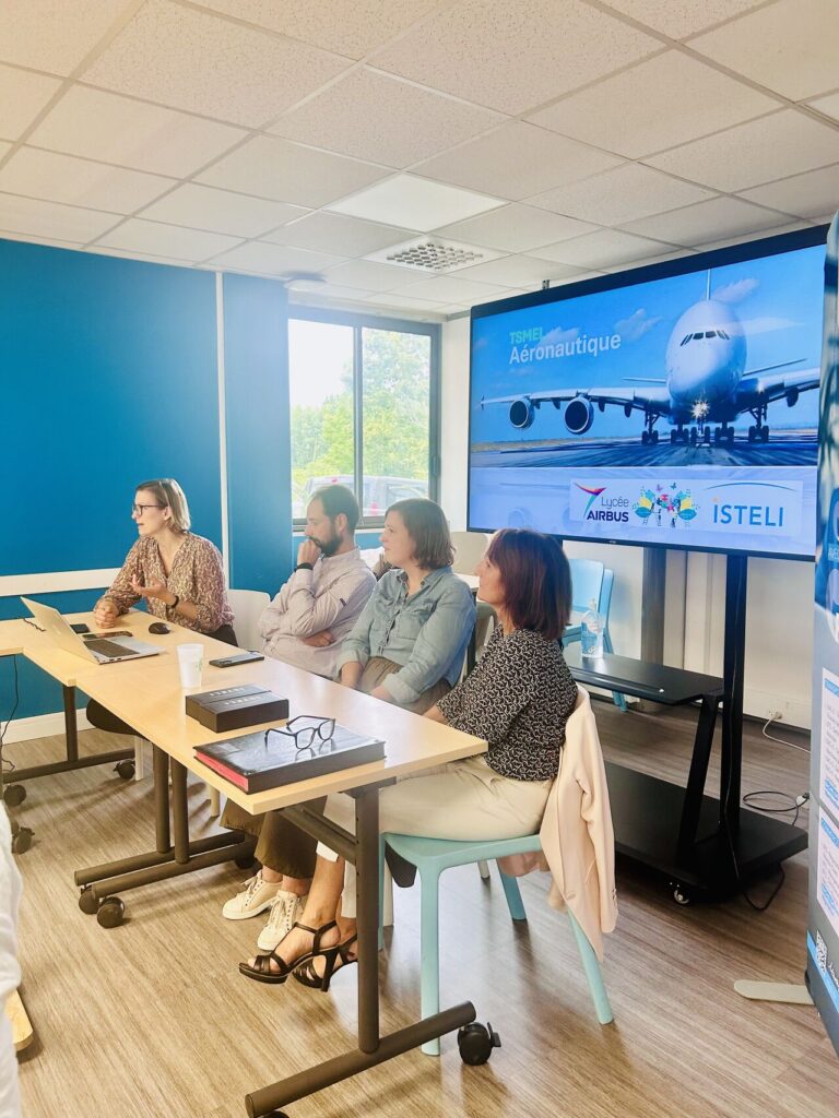 Une réunion est en cours avec plusieurs personnes assises devant un écran affichant un thème aéronautique dans une salle lumineuse.