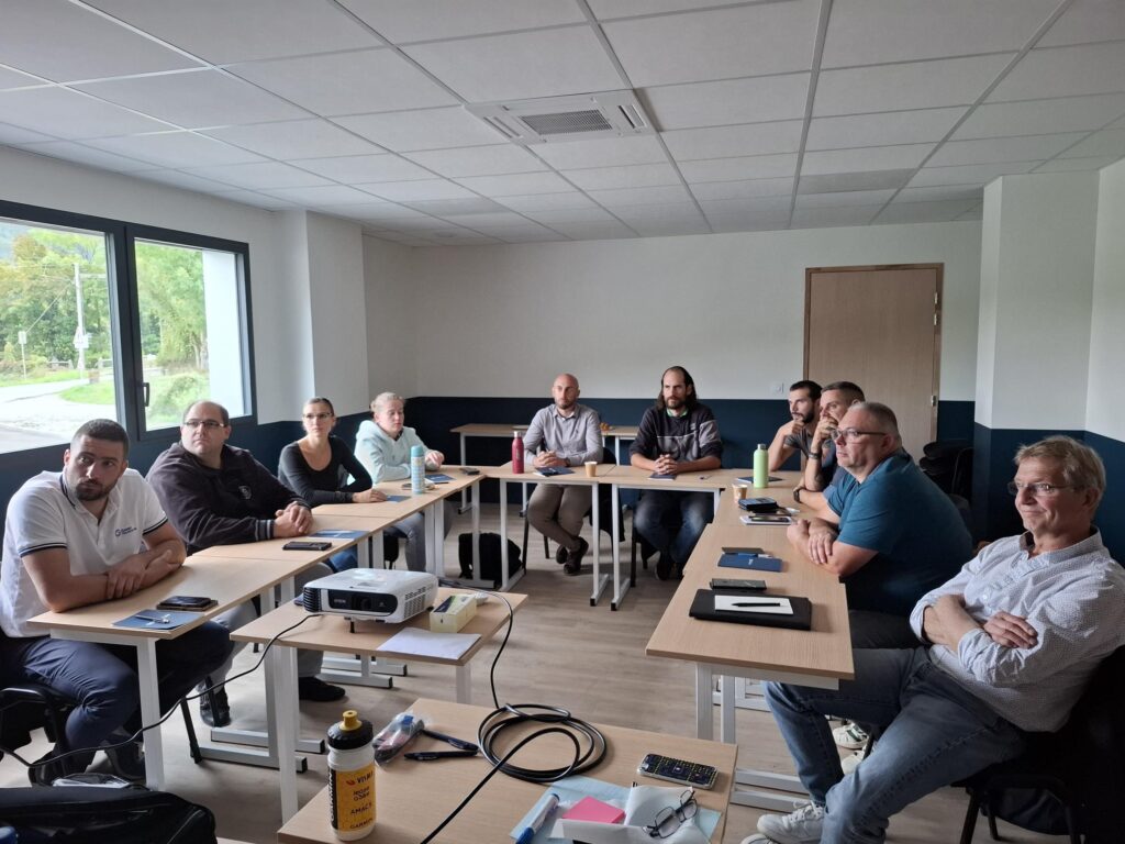 Une réunion se déroule dans une salle avec plusieurs personnes assises autour de tables, en attentant un présentateur.
