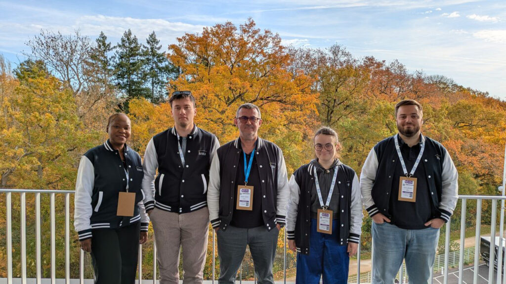Un groupe de cinq personnes, vêtues de vestes noires et blanches, pose sur un balcon avec des arbres aux feuilles d'automne en arrière-plan.
