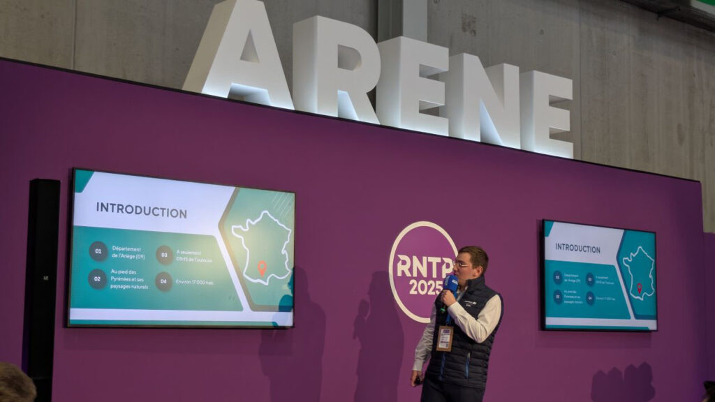 Un présentateur discute devant un écran montrant une carte de la France et des informations sur le département de l'Ariège lors d'un événement intitulé RNTP 2025.