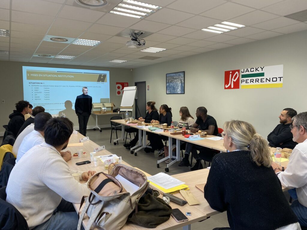Une salle de formation où un formateur présente à un groupe d'apprenants assis autour d'une table, avec des documents et de la nourriture visible.