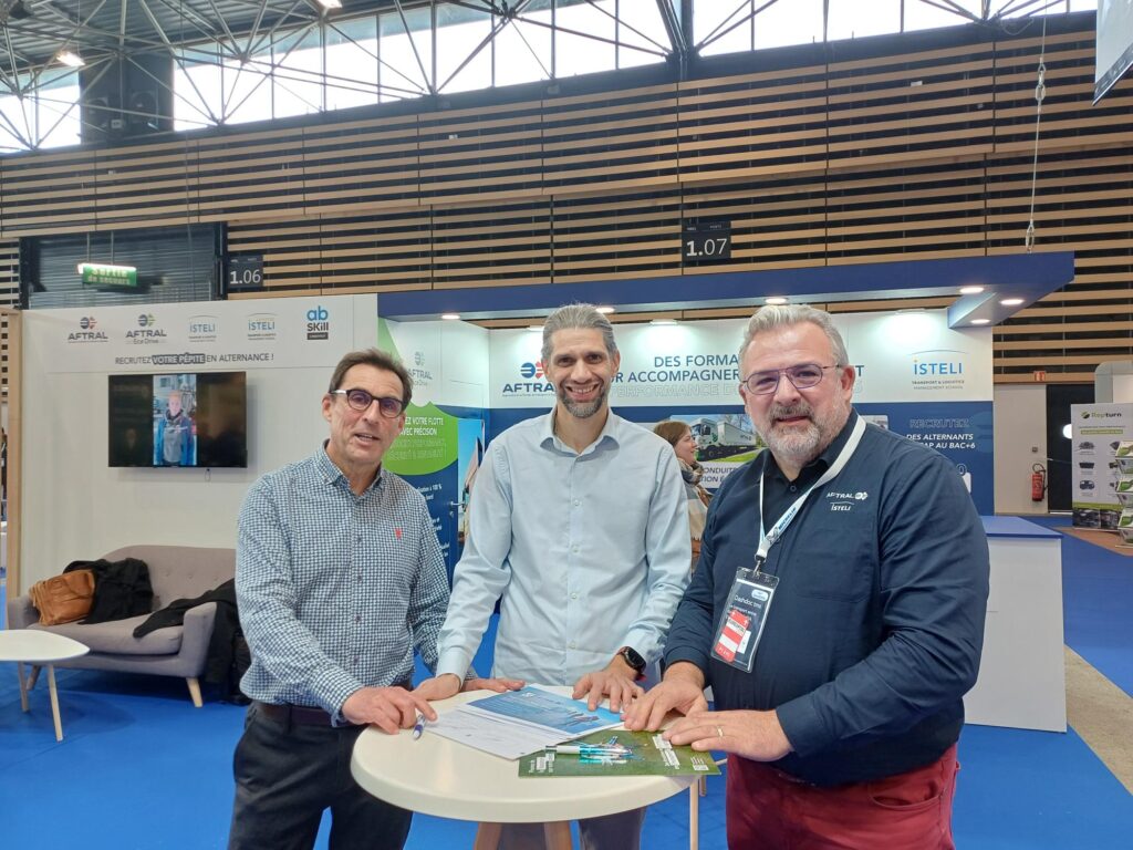 Trois hommes posent ensemble, souriants, près d'un stand lors d'un événement, avec un document sur une table entre eux.