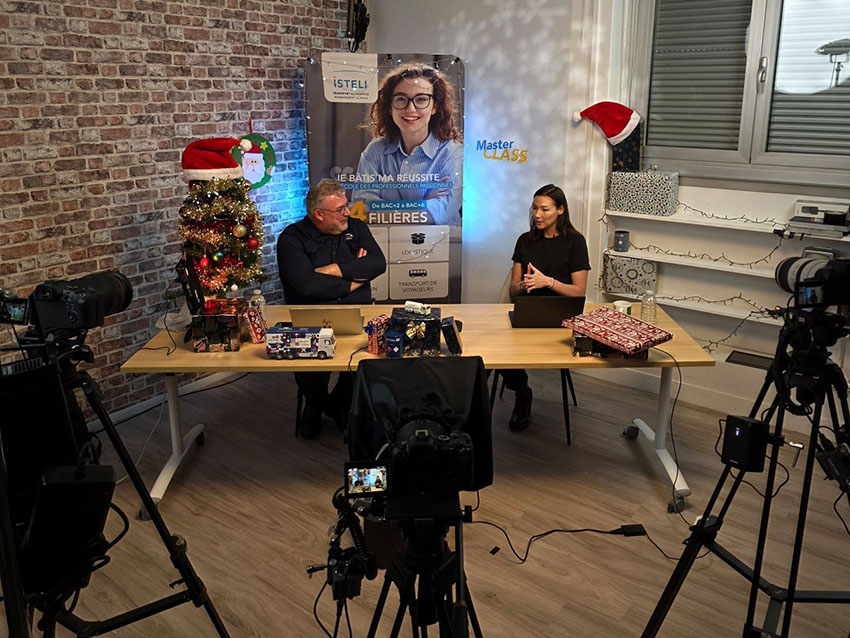 Une scène de tournage avec deux personnes assises à une table décorée pour les fêtes, entourées de caméras et d'un arbre de Noël.