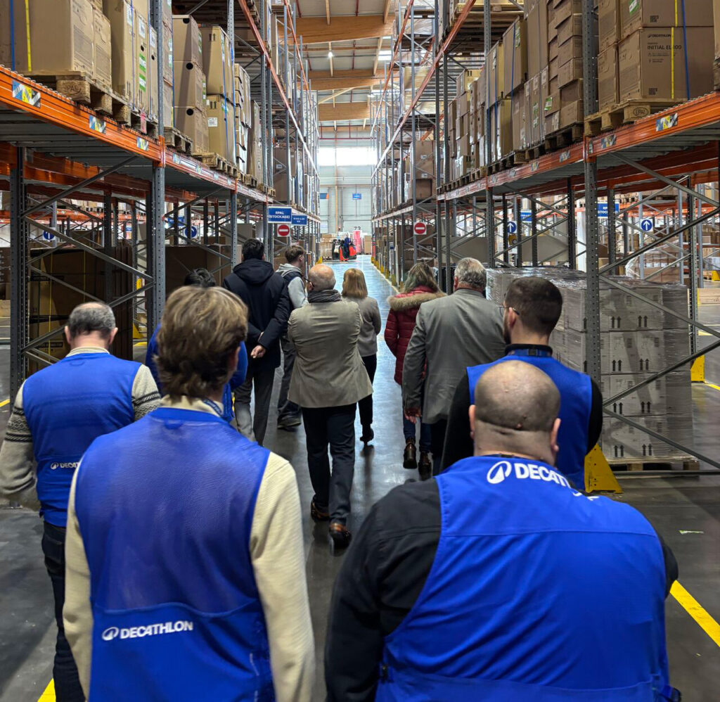 Un groupe de personnes portant des gilets bleus marche dans un entrepôt avec des étagères remplies de cartons.