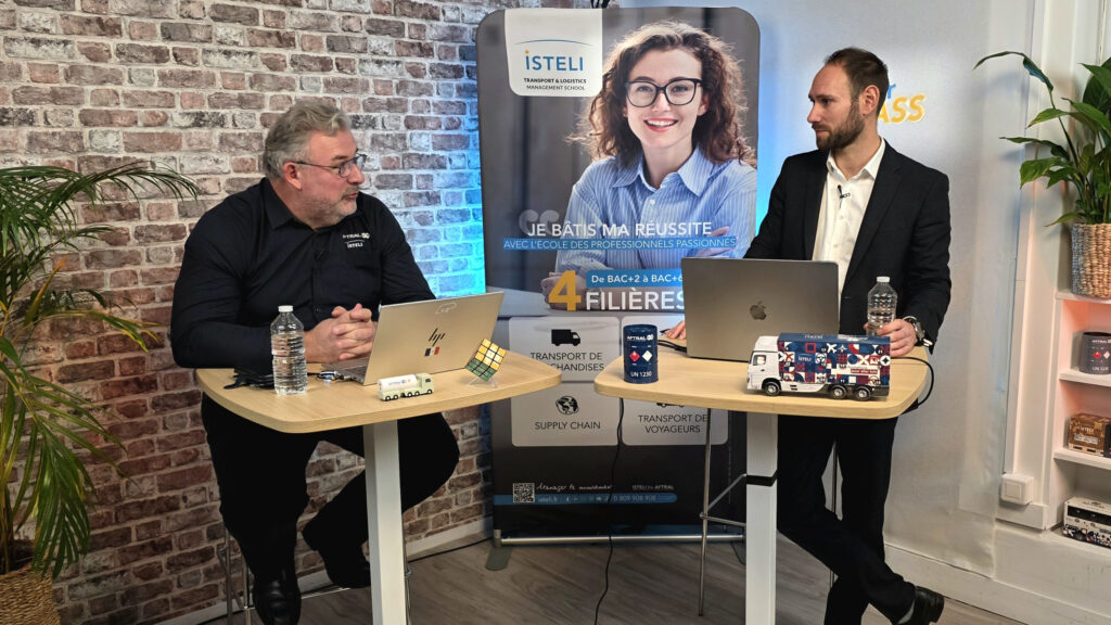 Deux hommes discutent devant des ordinateurs portables et un panneau de l'école ISTELI, avec des accessoires liés à la logistique sur une table.