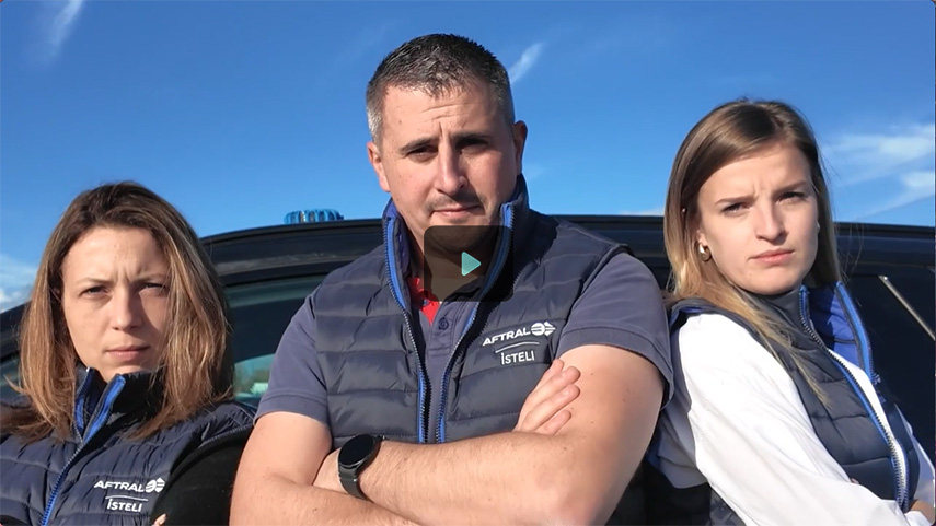 Trois personnes, aux expressions sérieuses, se tiennent devant un véhicule, avec un ciel bleu en arrière-plan.