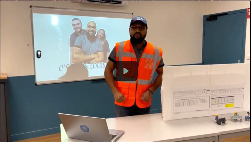 Un présentateur portant un gilet fluorescent se tient devant un tableau avec des graphiques et un ordinateur portable, tandis qu'une vidéo est projetée en arrière-plan.