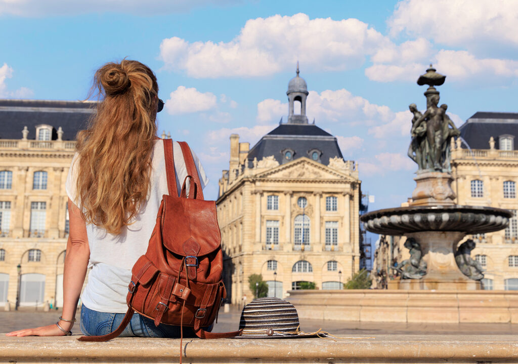 Une jeune femme aux cheveux longs et bouclés, portant un sac à dos en cuir et assise sur un mur, admire une élégante fontaine et des bâtiments historiques sous un ciel bleu parsemé de nuages.