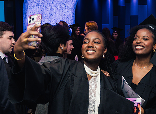 Deux jeunes femmes en tenue de diplôme prennent un selfie entourées de camarades de classe.
