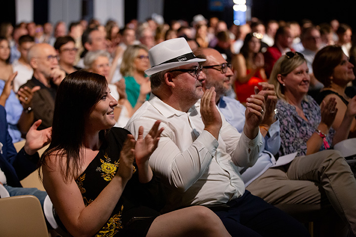 Une foule de spectateurs applaudit lors d'un événement, avec des personnes variées assises sur des chaises.