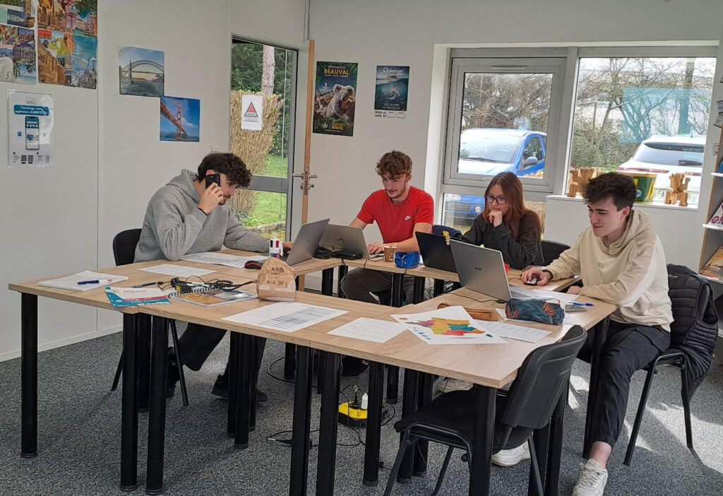 Quatre jeunes adultes travaillent ensemble autour d'une table, utilisant des ordinateurs portables et des documents, dans un environnement lumineux et moderne.