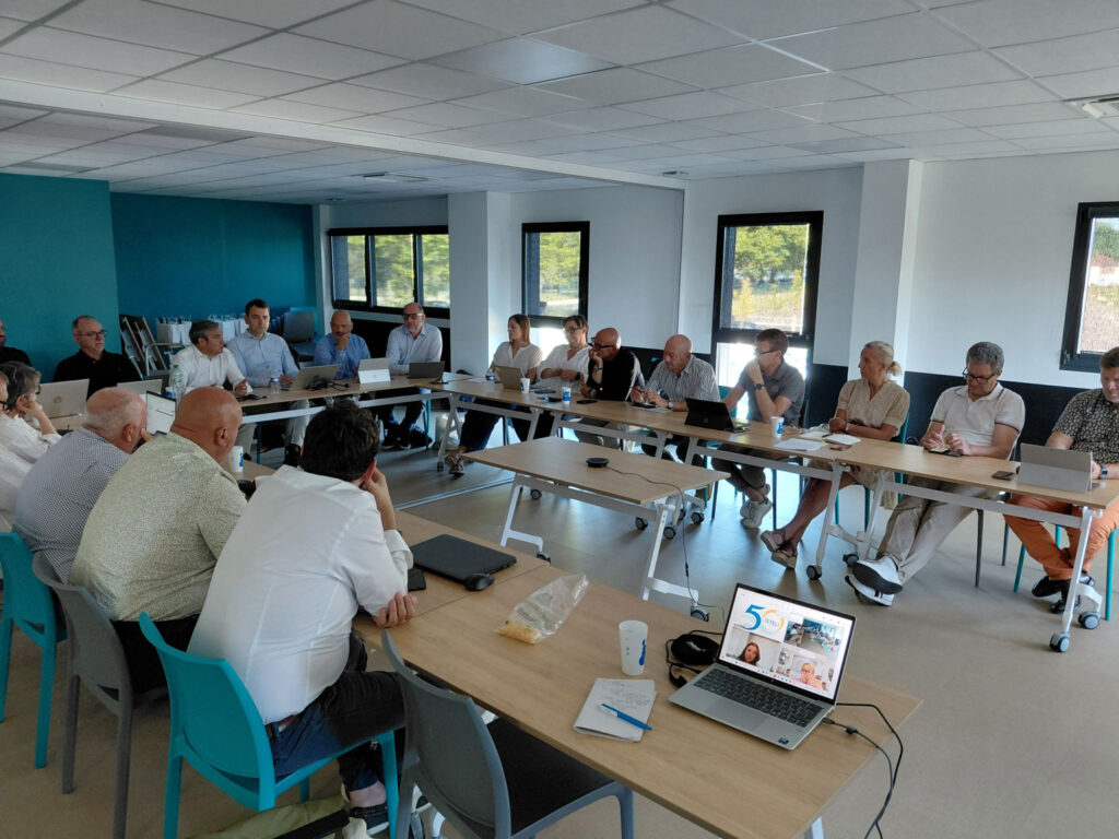 Une réunion se déroule dans une salle moderne avec des participants assis autour de tables, tous attentifs à un intervenant au centre.