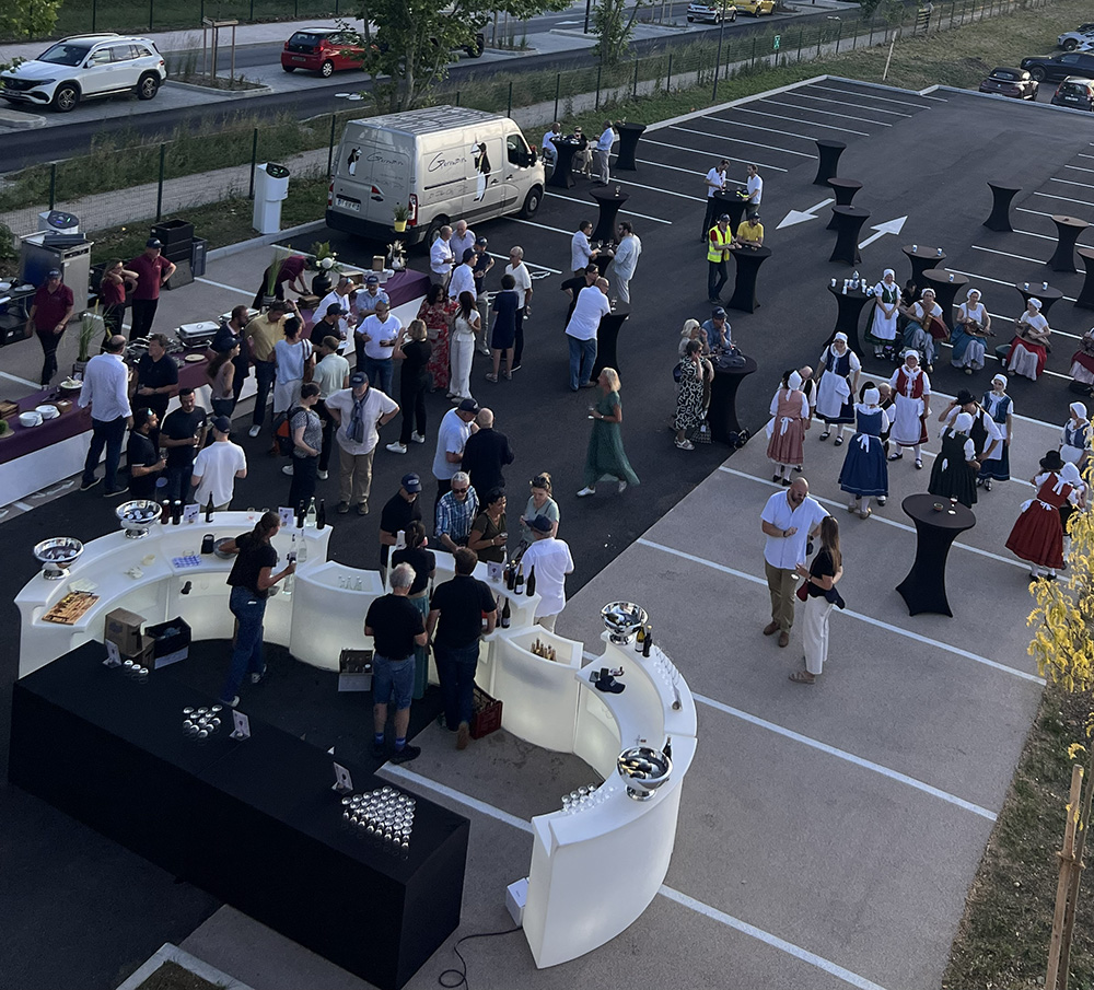 Un événement festif en plein air avec de nombreuses personnes conversant autour de tables, une ambiance animée et des stands de boissons dans un parking.