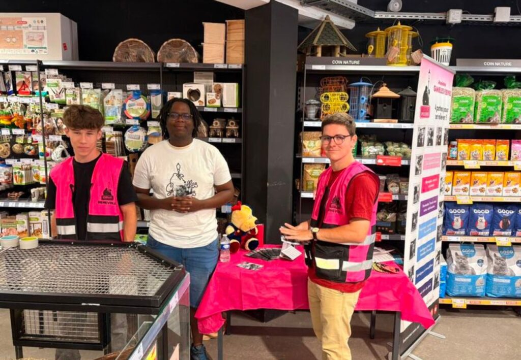 Trois personnes se tiennent dans un magasin, entourées de divers produits pour animaux, avec des tables et des étagères remplies d'articles.
