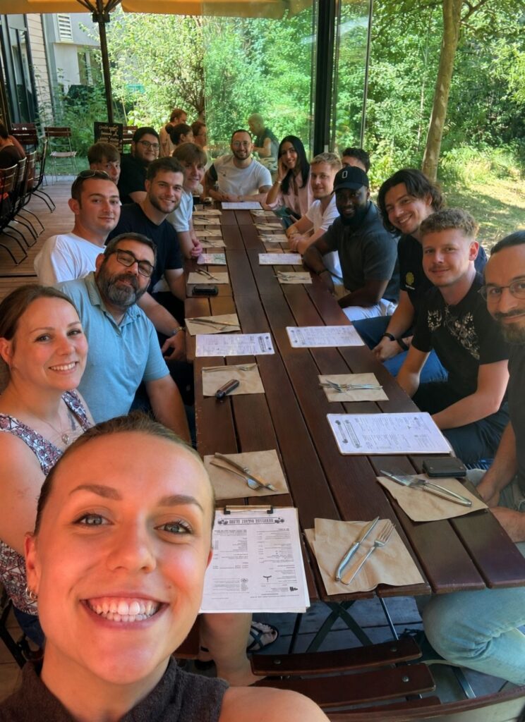 Un groupe de personnes souriantes pose ensemble autour d'une table dans un restaurant, avec des menus et des couverts devant eux, entourés de verdure.