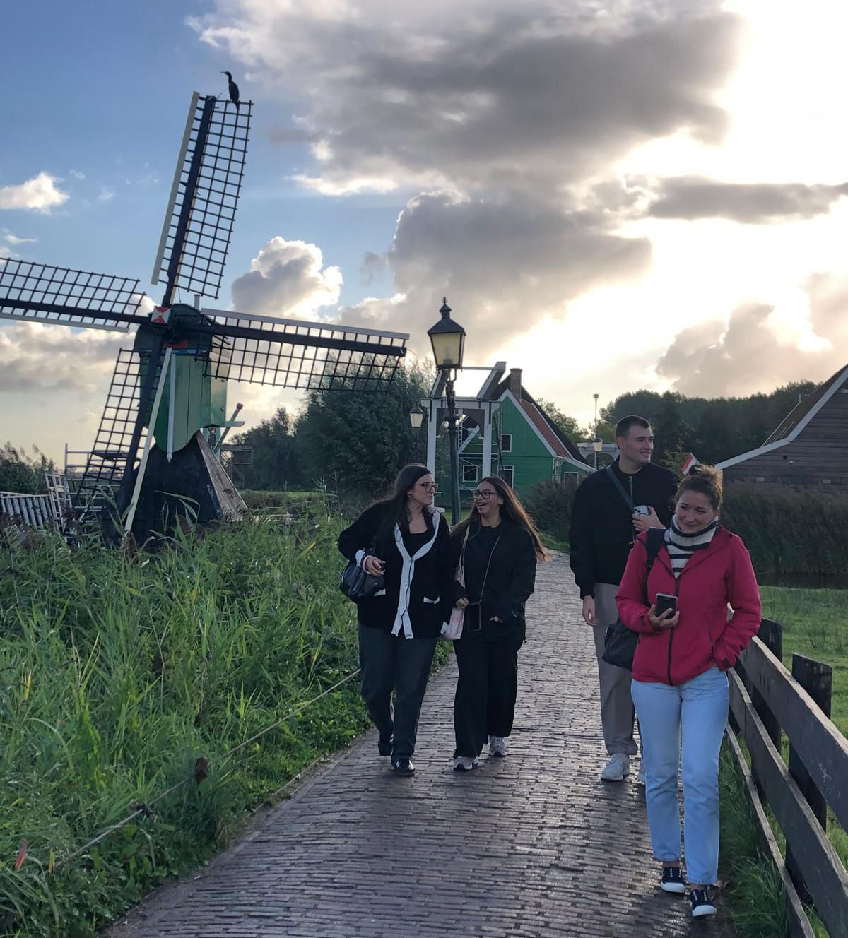 Un groupe de quatre personnes se promène sur un chemin en briques, avec un moulin à vent et des maisons colorées en arrière-plan sous un ciel nuageux.