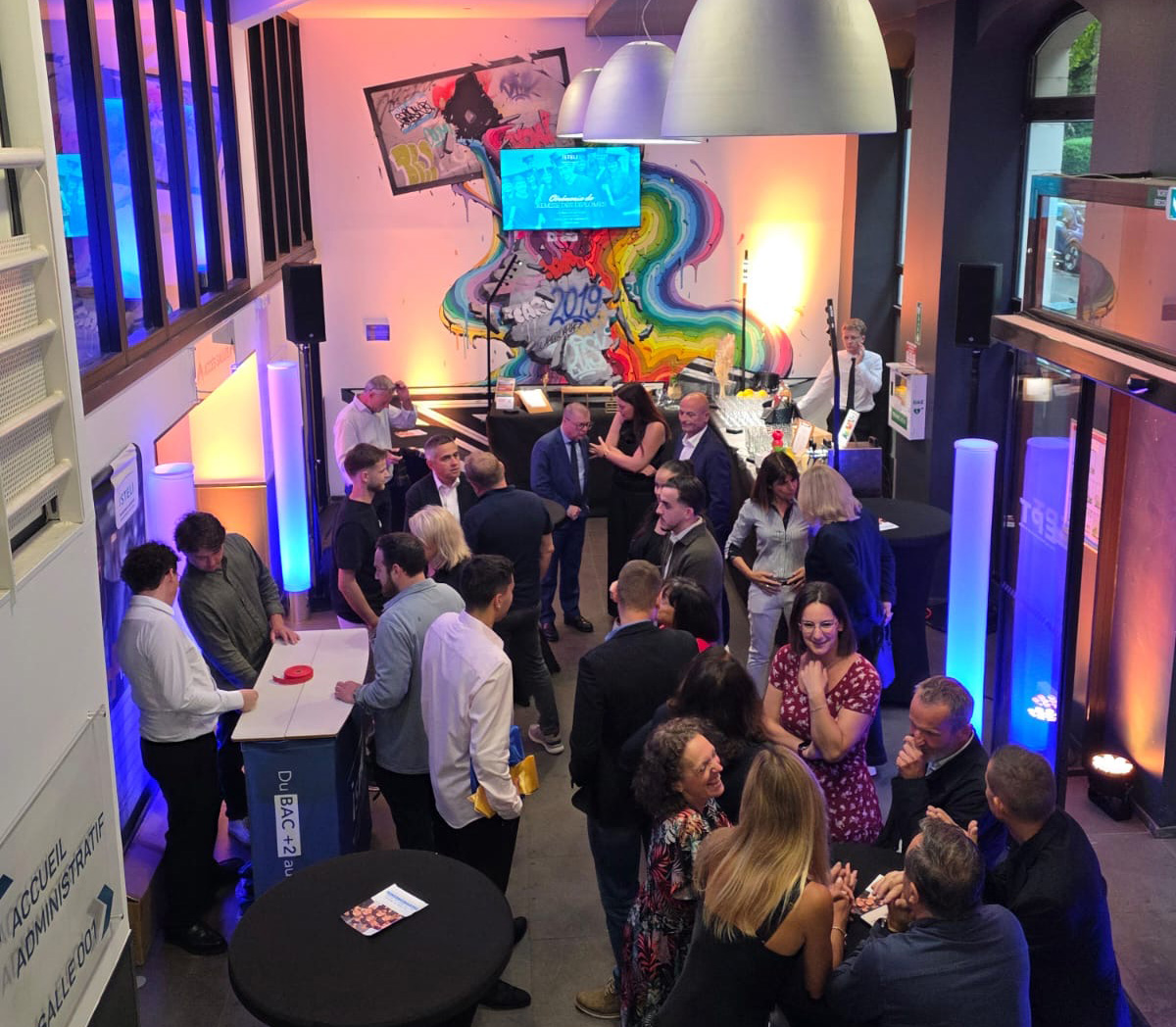 Une foule de personnes interagissent dans un espace élégant décoré avec des lumières colorées et un mur artistique, lors d'un événement social.