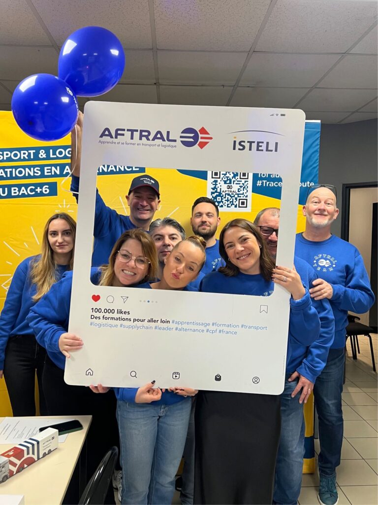 Un groupe de personnes en sweat-shirts bleus sourit et pose devant un panneau portant le logo d'AFTRAL et d'ISTELI, tenant des ballons et un cadre représentant un post Instagram.