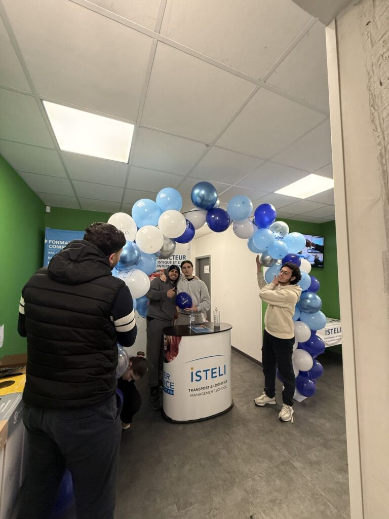 Des personnes se rassemblent sous un arc fait de ballons bleus et blancs dans un espace lié à une école de gestion.