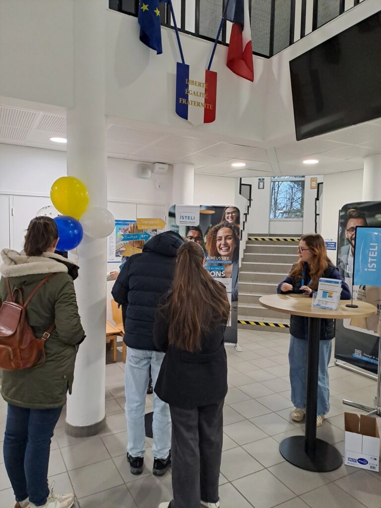 Un groupe de personnes discute dans un hall décoré avec des drapeaux et des ballons, tandis qu'une personne présente des informations à un stand.