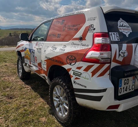 Un véhicule tout-terrain décoré avec des logos sponsorisés, stationné sur un terrain verdoyant sous un ciel nuageux.