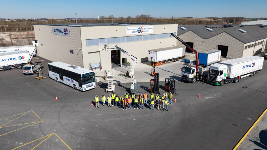 Une vue aérienne d'un centre de formation en transport avec des véhicules, des engins de levage et un groupe de personnes en gilets de sécurité.