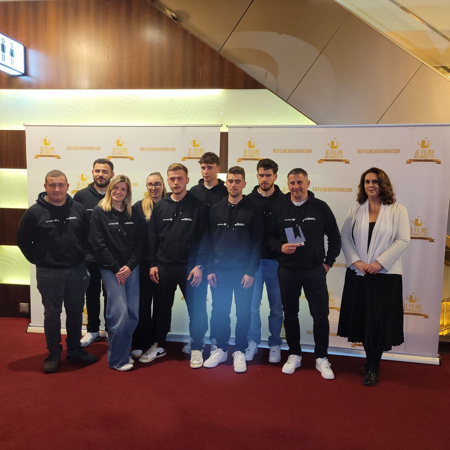 Un groupe de jeunes adultes en sweatshirts noirs se tient devant un fond avec des logos, probablement lors d'un événement ou d'une remise de prix.