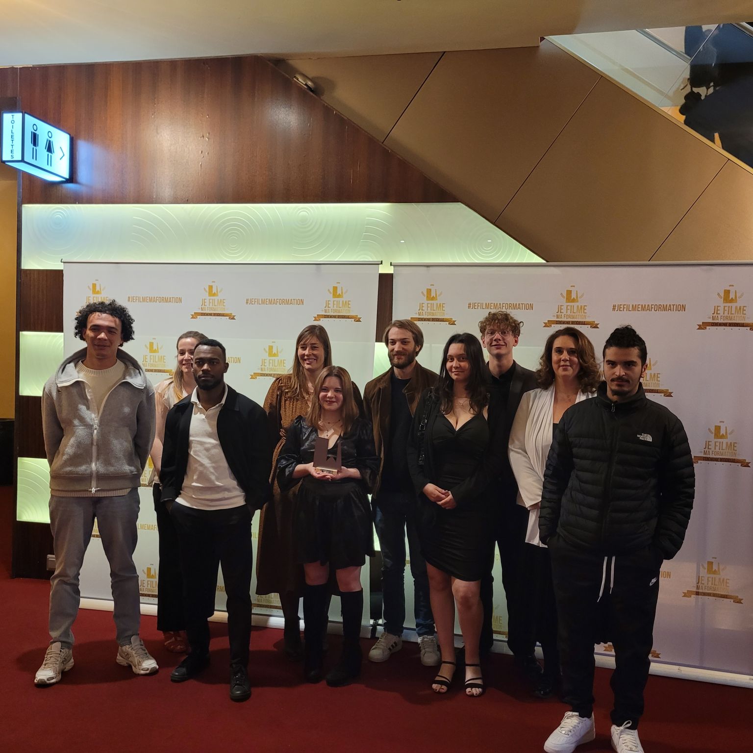 Un groupe de personnes se tient en pose souriante devant un panneau sur lequel est écrit "JE FILME" dans une ambiance de soirée.