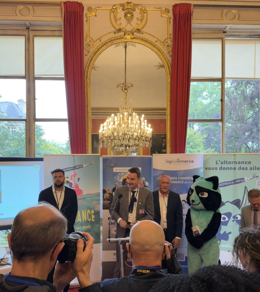 Une scène de conférence avec plusieurs intervenants, dont un homme en costume devant un podium, accompagné d'une mascotte en costume d'animal et des affiches promotionnelles en arrière-plan.