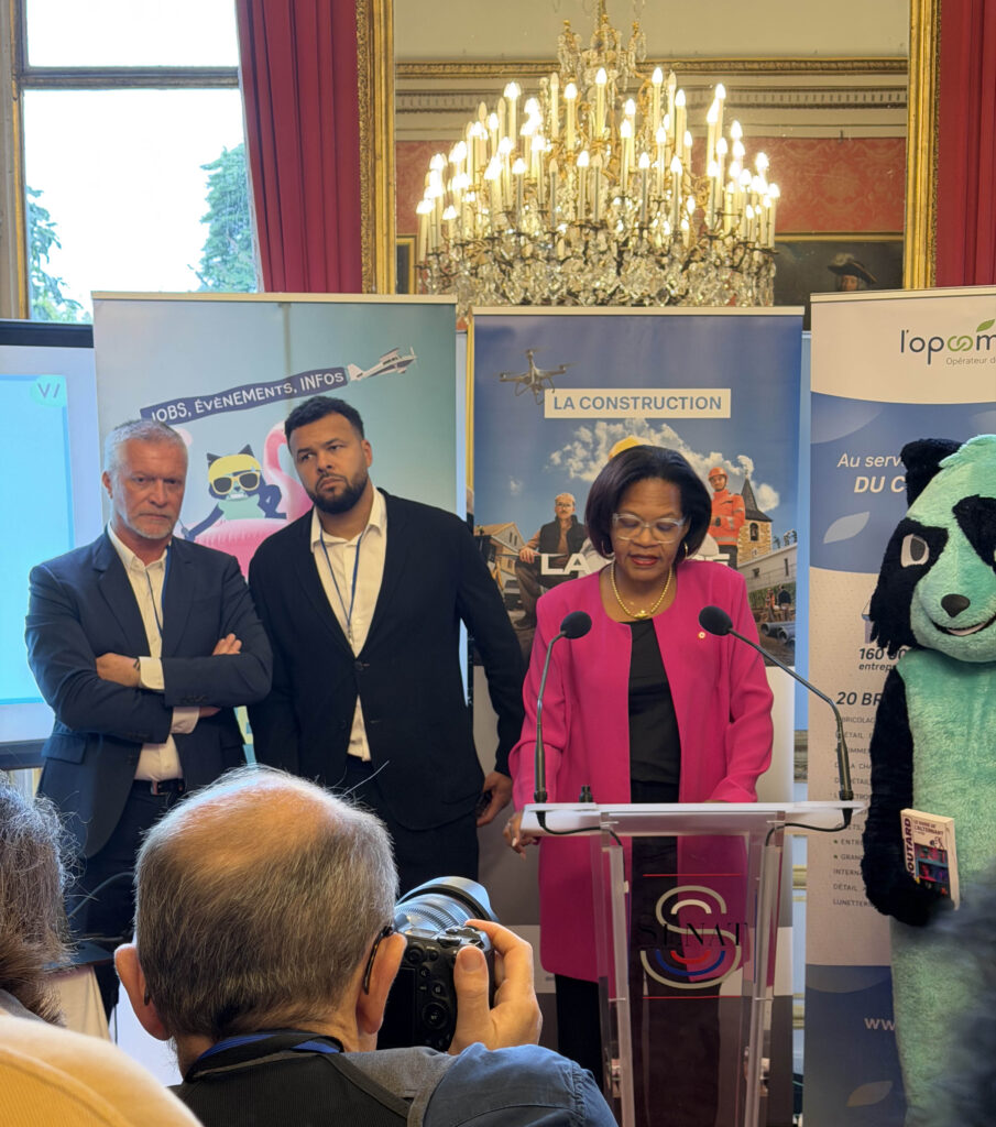 Une femme en costume rose se tient derrière un pupitre de conférence, entourée de deux hommes en costumes sombres et d'un personnage en costume de panda, lors d'un événement dans une salle ornée de lustres.