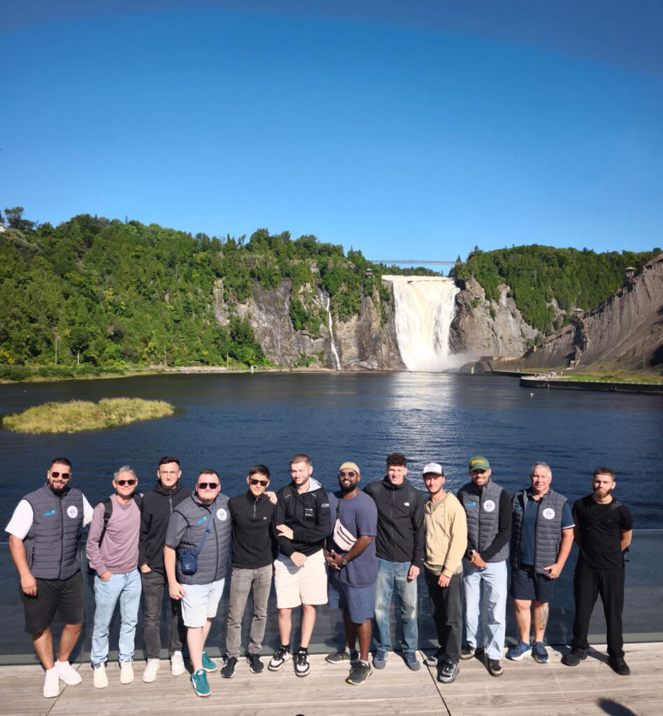 Un groupe d'hommes pose en avant-plan avec une spectaculaire cascade en arrière-plan, sous un ciel bleu clair.